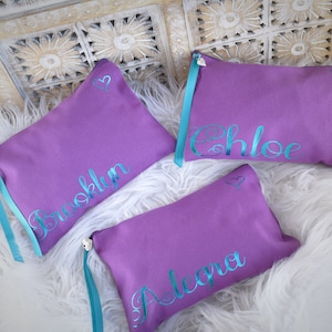May include: Three purple zippered pouches with teal ribbon ties and silver heart charms. Each pouch has a name embroidered in teal lettering: Brooklyn, Chloe, and Alecia.