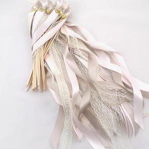 May include: A bundle of ivory and blush pink satin and lace ribbon wands with gold bells at the top. The wands are tied together with a blush pink ribbon.