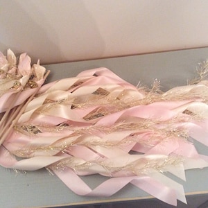 May include: A set of pink and ivory ribbon wands with gold glitter and bells. The wands are tied together and ready for a celebration.