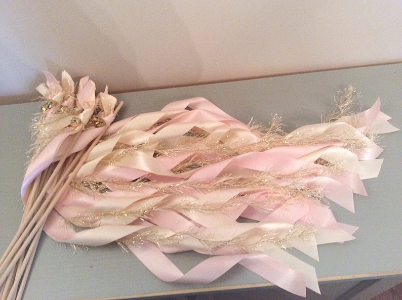 May include: A set of pink and ivory ribbon wands with gold glitter and bells. The wands are tied together and ready for a celebration.