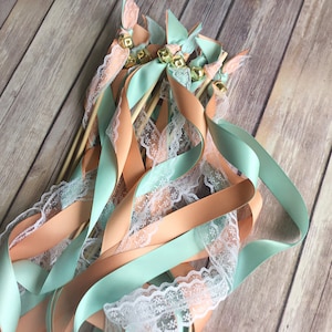 May include: A bundle of wedding ribbon wands with peach and mint green satin ribbons and white lace. The wands have wooden sticks and gold bells.
