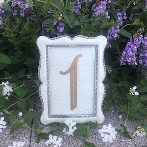 May include: A white frame with a silver border and a white background. The number "1" is written in gold glitter inside the frame.