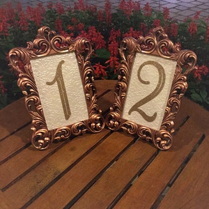 May include: Two ornate gold-colored frames with a white background and gold glitter numbers 1 and 2. The frames are on a wooden table.