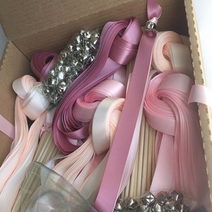 May include: A box filled with pink and peach satin ribbon wands. The wands are made with wooden dowels and have silver bells attached to the ends. The box also contains extra bells and ribbon.