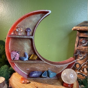 May include: A crescent moon-shaped shelf made of wood with a red, brown, and gold finish. The shelf has three tiers and is filled with crystals, a skull, and a mushroom. A clear crystal ball sits on a wooden stand in front of the shelf.