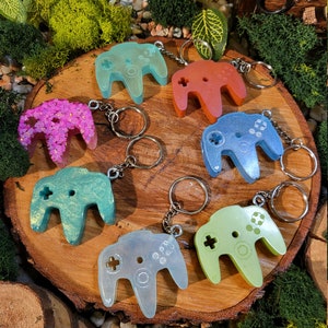 Puede incluir: Un conjunto de ocho llaveros de mando de Nintendo 64 de colores. Los llaveros están hechos de resina y presentan diferentes colores y detalles de purpurina.