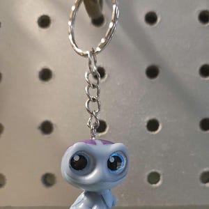 Pu&ograve; includere: Un portachiavi con una piccola statuetta di tartaruga in plastica azzurra con grandi occhi scuri e un motivo a guscio viola. La statuetta &egrave; fissata a un anello portachiavi in metallo argentato tramite una corta catena a tre maglie.