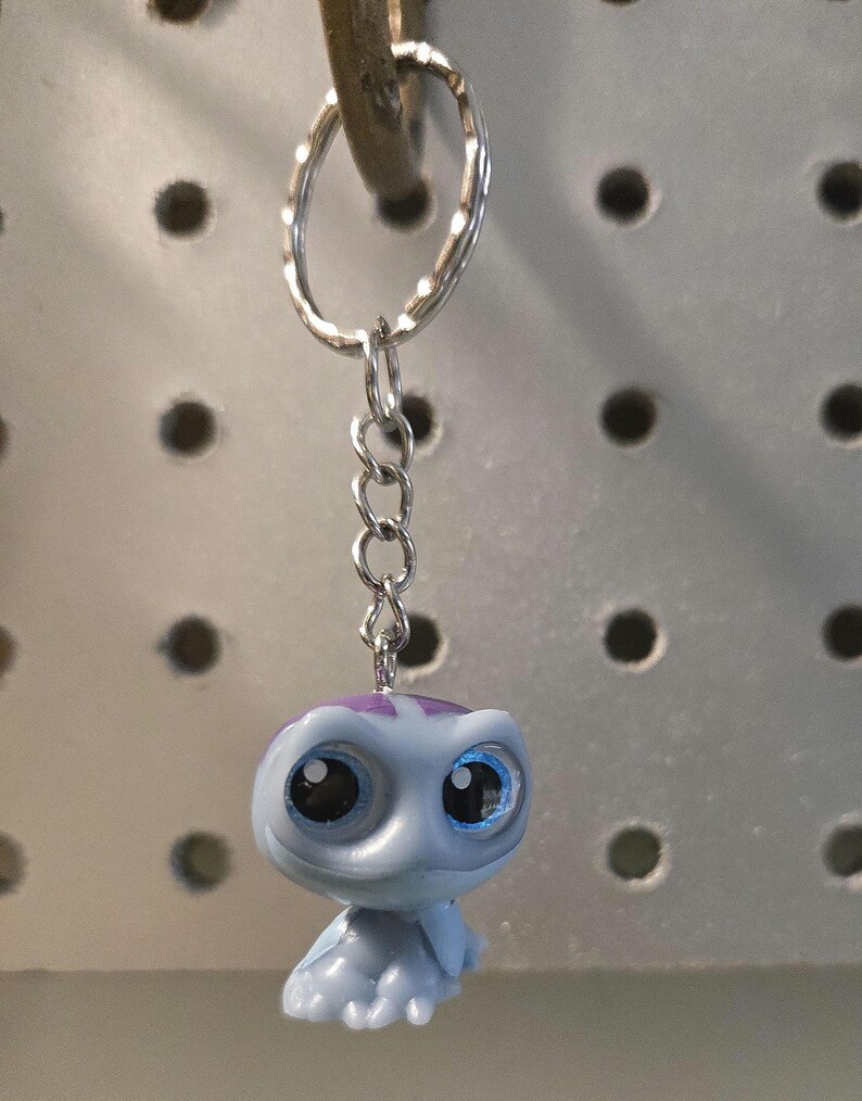 Pu&ograve; includere: Un portachiavi con una piccola statuetta di tartaruga in plastica azzurra con grandi occhi scuri e un motivo a guscio viola. La statuetta &egrave; fissata a un anello portachiavi in metallo argentato tramite una corta catena a tre maglie.