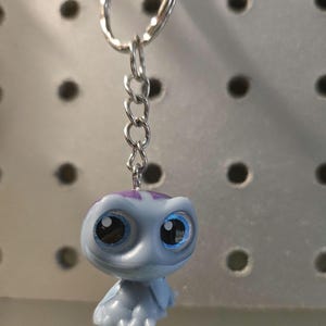 Pu&ograve; includere: Un portachiavi con una piccola statuetta di tartaruga azzurra con grandi occhi scuri e un segno viola sulla testa. La tartaruga &egrave; fissata a un anello diviso in metallo argentato tramite una corta catena a maglie in metallo argentato.