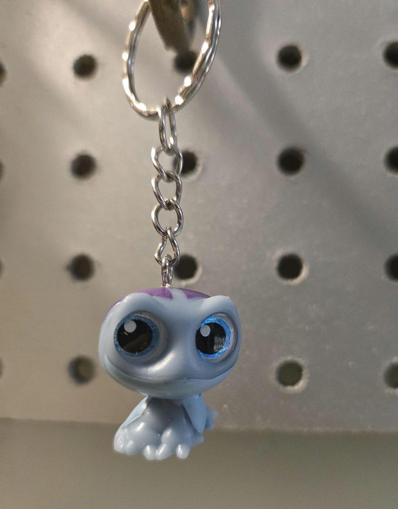 Pu&ograve; includere: Un portachiavi con una piccola statuetta di tartaruga azzurra con grandi occhi scuri e un segno viola sulla testa. La tartaruga &egrave; fissata a un anello diviso in metallo argentato tramite una corta catena a maglie in metallo argentato.