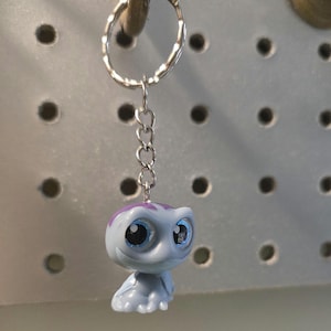 Pu&ograve; includere: Un portachiavi con una piccola statuetta di creatura azzurra con grandi occhi blu espressivi e una macchia viola sulla testa. La statuetta &egrave; fissata a un anello in metallo argentato tramite una corta catenella in metallo a quattro maglie.