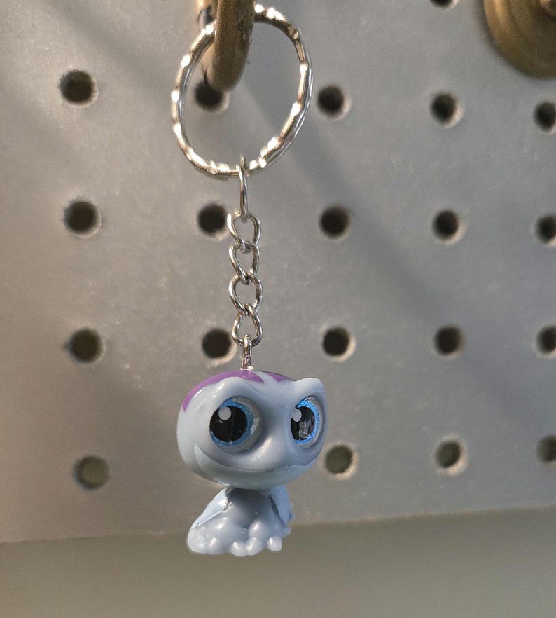 Pu&ograve; includere: Un portachiavi con una piccola statuetta di creatura azzurra con grandi occhi blu espressivi e una macchia viola sulla testa. La statuetta &egrave; fissata a un anello in metallo argentato tramite una corta catenella in metallo a quattro maglie.