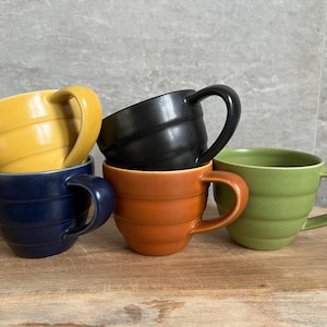 Può includere: Una pila di cinque tazze in ceramica di vari colori: giallo, nero, blu navy, arancione e verde. Ogni tazza ha una forma arrotondata, un manico e dettagli orizzontali a coste. Le tazze sono disposte su una superficie di legno.