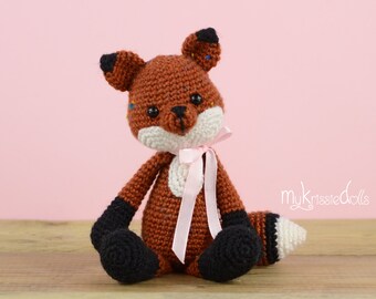 May include: A handmade crochet fox doll with brown, black, and white details. The fox has a white chest, a pink ribbon, and black paws. The doll is sitting on a wooden surface with a pink background.