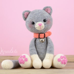 May include: A handmade crochet cat doll in gray, white, and pink. The cat has a pink nose, blush, and paw pads. It wears an orange collar with a silver bell. The doll is sitting with its legs and tail extended.