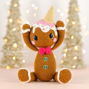 Op de afbeelding: Een handgemaakte gehaakte peperkoekenmanpop met een roze strik, een kegelvormige hoed en een roze en wit glazuurontwerp. De pop heeft zwarte ogen en zit met de armen omhoog. De voeten en handen van de pop hebben een witte rand.