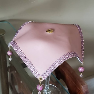 Puede incluir: Una cubierta de copa de vino de tela rosa con un botón dorado y flecos de cuentas rosas.