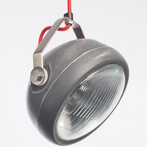 Puede incluir: Una lámpara colgante de metal gris con una lente de vidrio transparente y un cable rojo. La lámpara tiene un acabado desgastado y un diseño sencillo.