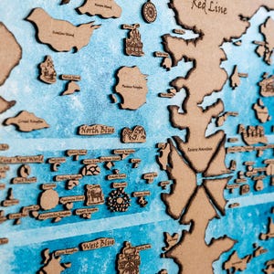 Puede incluir: Un mapa de madera con una combinación de colores azul y marrón. El mapa presenta recortes detallados de islas y continentes, con etiquetas de texto como "Red Line", "North Blue" y "West Blue". El mapa está enmarcado en madera oscura.