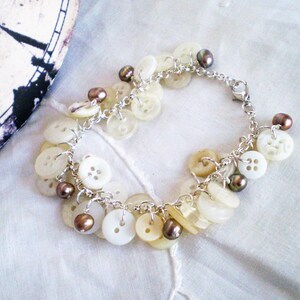 May include: A handmade bracelet featuring a collection of ivory-colored buttons and small, dark brown beads. The buttons vary in size and style, attached to a silver-toned chain. The bracelet is displayed on a white, textured surface.