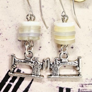 May include: A pair of silver sewing machine earrings with white and grey spools hanging from them. The earrings are hanging from a clock face.