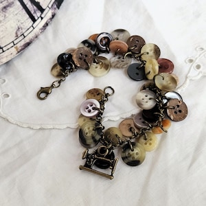 May include: A brown and gold bracelet featuring a variety of buttons and a sewing machine charm. The bracelet is made of metal and has a clasp fastening.