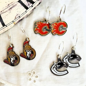 May include: A set of six silver earrings featuring different sports team logos. The logos include the Calgary Flames, the Ottawa Senators, and the Vancouver Canucks.