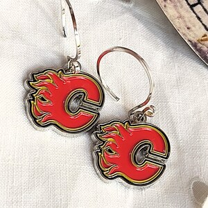 May include: A pair of silver hoop earrings with a red and yellow Calgary Flames logo charm. The logo features a stylized "C" with flames on either side.
