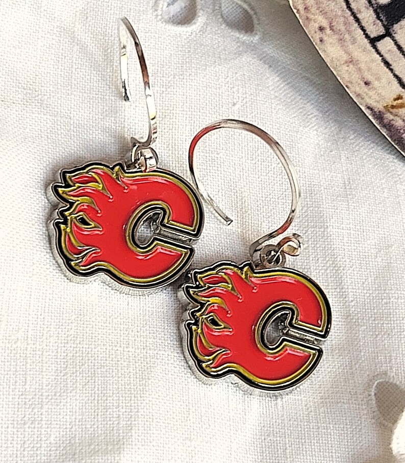 May include: A pair of silver hoop earrings with a red and yellow Calgary Flames logo charm. The logo features a stylized "C" with flames on either side.