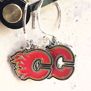 May include: A pair of silver earrings with a red and yellow Calgary Flames logo. The logo is a stylized "C" with flames on the side.