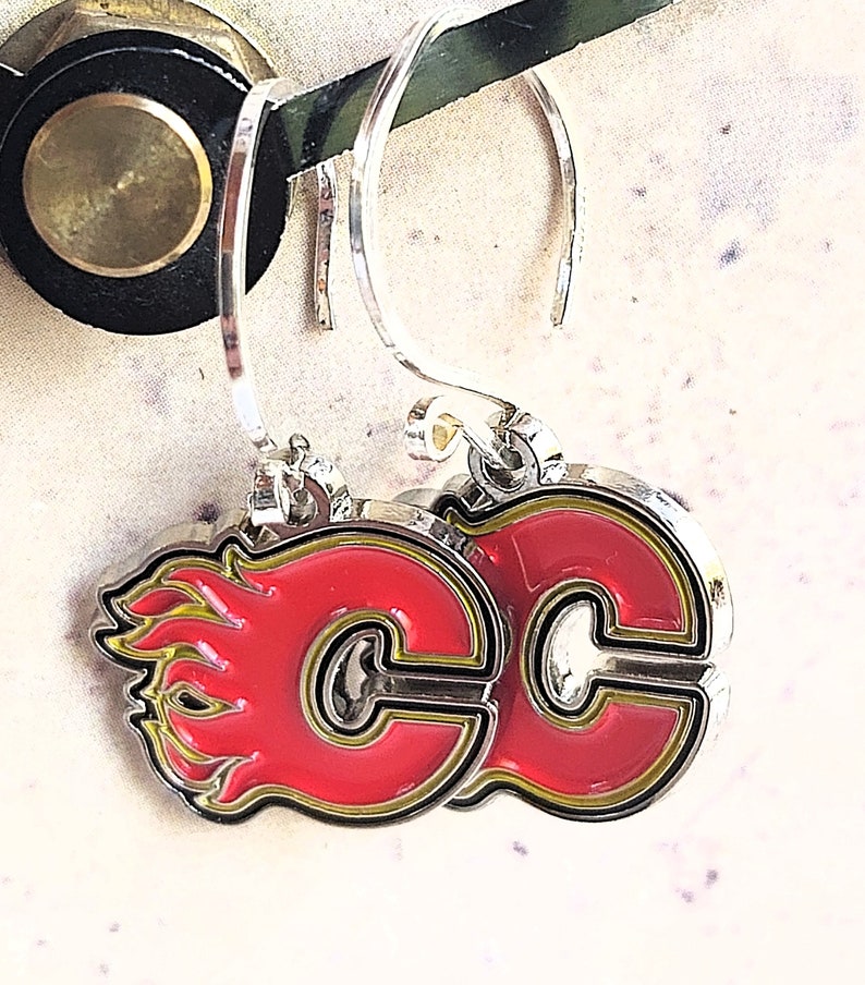 May include: A pair of silver earrings with a red and yellow Calgary Flames logo. The logo is a stylized "C" with flames on the side.