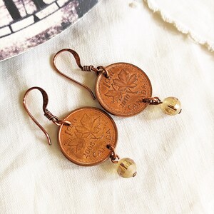 Peut inclure: Une paire de boucles d'oreilles en cuivre avec un sou canadien orn&eacute; d'une feuille d'&eacute;rable et une perle de verre transparente. Le sou est dat&eacute; de 2008.