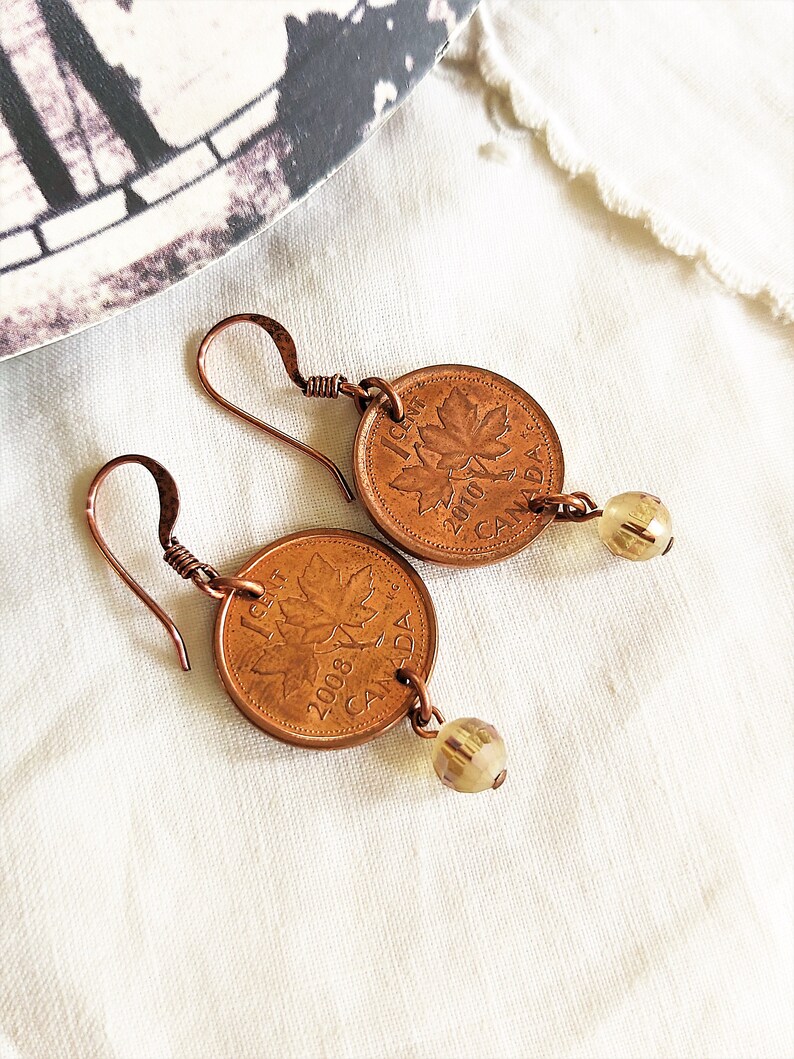 Peut inclure: Une paire de boucles d'oreilles en cuivre avec un sou canadien orn&eacute; d'une feuille d'&eacute;rable et une perle de verre transparente. Le sou est dat&eacute; de 2008.