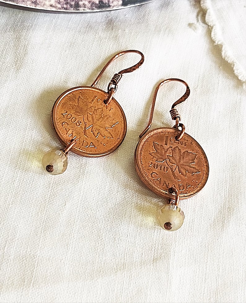 Peut inclure: Une paire de boucles d'oreilles en fil de cuivre et des pi&egrave;ces de monnaie canadiennes d'un cent de 2008 et 2010. Chaque pi&egrave;ce pr&eacute;sente un motif de feuille d'&eacute;rable et le mot "CANADA". Les boucles d'oreilles sont rehauss&eacute;es de petites perles de verre transparentes.