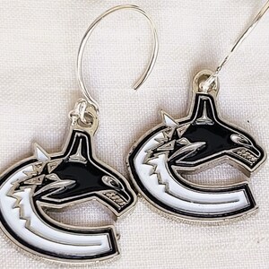 May include: A pair of silver earrings with a black and white hockey team logo. The logo features a stylized orca whale with a white 'C' shape behind it.