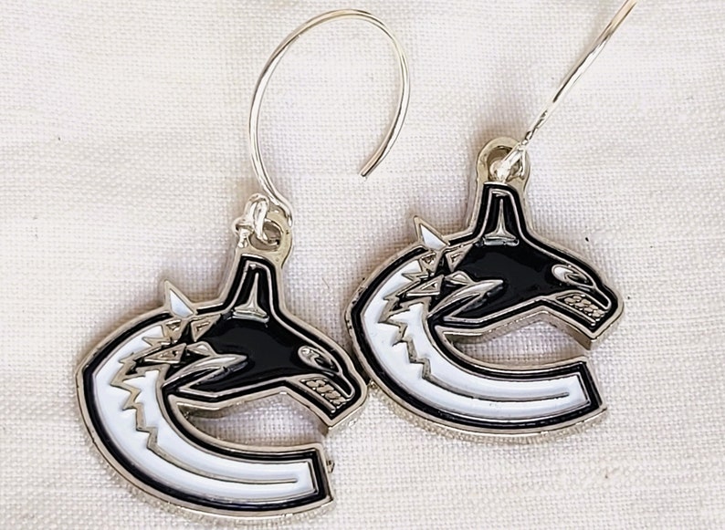 May include: A pair of silver earrings with a black and white hockey team logo. The logo features a stylized orca whale with a white 'C' shape behind it.