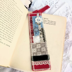 May include: A handmade fabric bookmark with a patchwork design. The bookmark is made of various fabrics in shades of red, blue, white, and gray. The bookmark is decorated with lace and a small white flower.