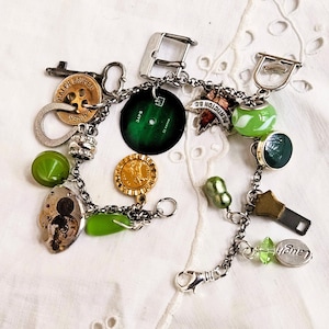 May include: A silver charm bracelet with various charms, including a green vinyl record, a gold coin, and a charm that says "Laugh". Other charms include green beads, a key, and a watch buckle. The bracelet is on a white, textured surface.