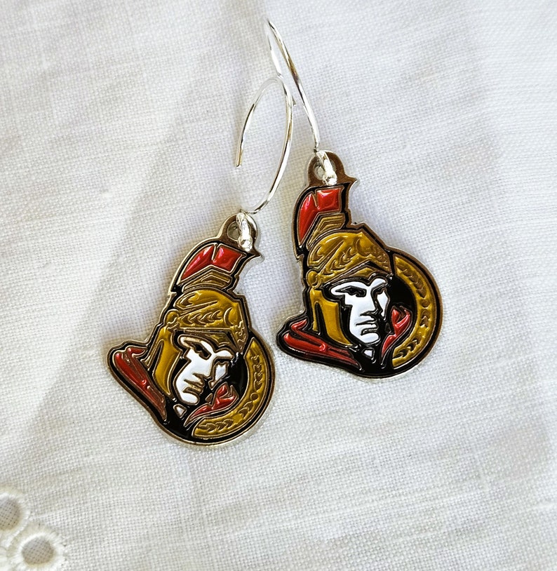 May include: A pair of silver earrings with a gold and red Ottawa Senators hockey team logo charm. The logo features a Roman soldier's head with a helmet and a wreath.