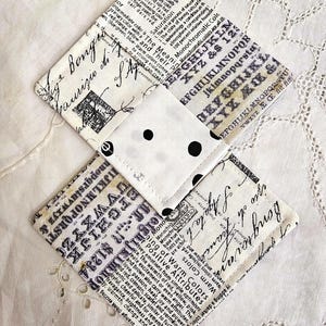 May include: Three fabric coasters with a vintage newspaper print design. The coasters have black text and graphics on a cream background, with a small polka dot square in the centre. The coasters are made of fabric.