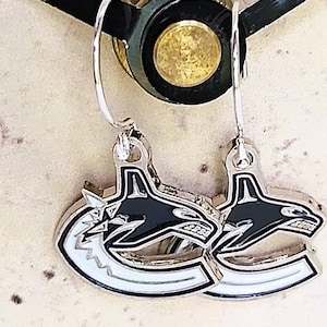 May include: A pair of silver earrings with a black and white orca design. The orca is facing right and has a white outline. The earrings are hanging from silver hooks.