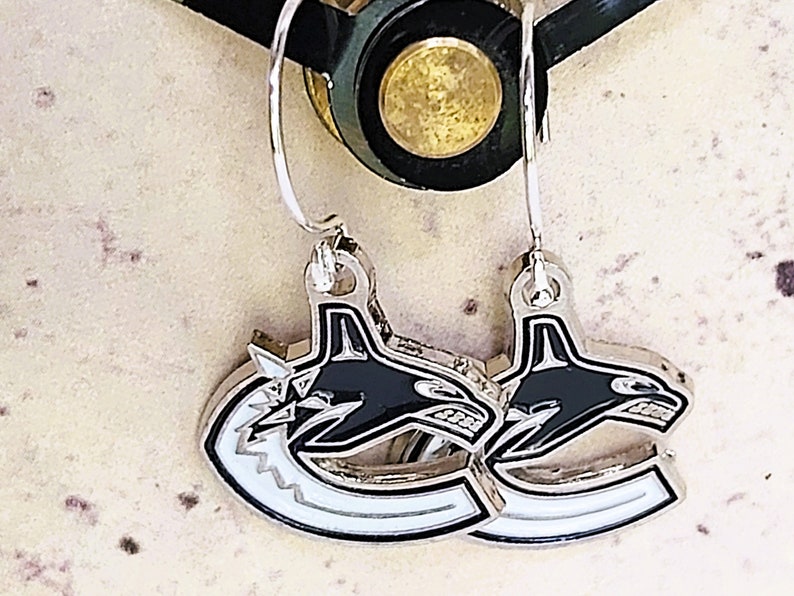 May include: A pair of silver earrings with a black and white orca design. The orca is facing right and has a white outline. The earrings are hanging from silver hooks.