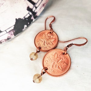 Peut inclure: Une paire de boucles d'oreilles en pi&egrave;ces de monnaie canadiennes d'un cent en cuivre de 2004, chacune avec un motif de feuille d'&eacute;rable. Les pi&egrave;ces sont attach&eacute;es &agrave; des perles de verre transparentes et &agrave; un fil de cuivre.