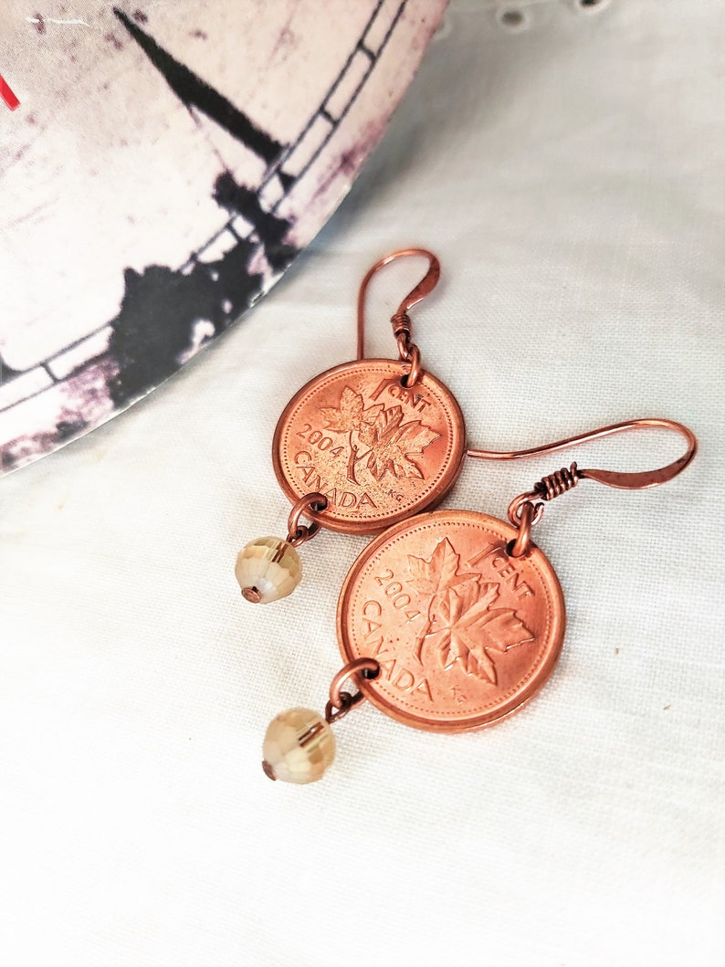 Peut inclure: Une paire de boucles d'oreilles en pi&egrave;ces de monnaie canadiennes d'un cent en cuivre de 2004, chacune avec un motif de feuille d'&eacute;rable. Les pi&egrave;ces sont attach&eacute;es &agrave; des perles de verre transparentes et &agrave; un fil de cuivre.