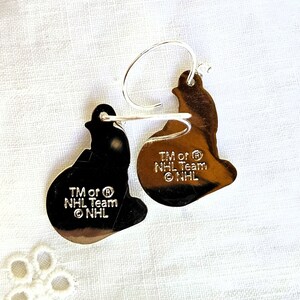 May include: A pair of silver earrings shaped like hockey pucks. The earrings have the NHL logo and the text "TM or &reg; NHL Team &copy; NHL".