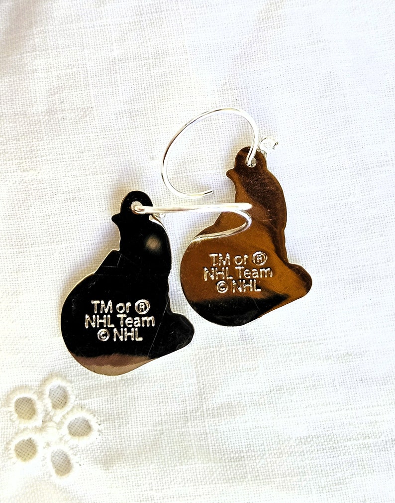 May include: A pair of silver earrings shaped like hockey pucks. The earrings have the NHL logo and the text "TM or &reg; NHL Team &copy; NHL".