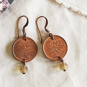 Peut inclure: Une paire de boucles d'oreilles en cuivre avec des pi&egrave;ces de monnaie canadiennes de 1 cent de 2008 et 2010, chacune avec un motif de feuille d'&eacute;rable et une petite perle ambr&eacute;e qui pend en dessous.