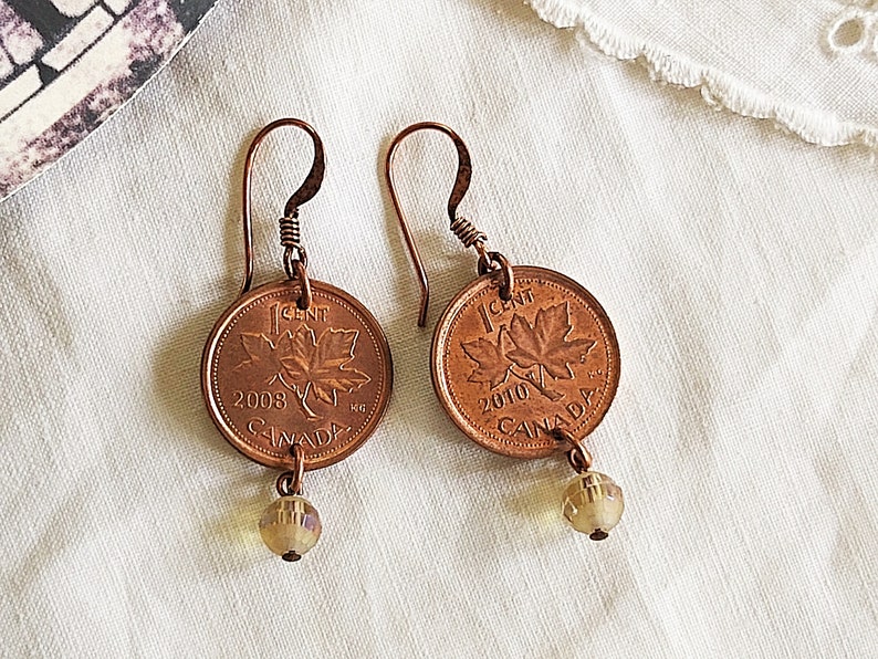 Peut inclure: Une paire de boucles d'oreilles en cuivre avec des pi&egrave;ces de monnaie canadiennes de 1 cent de 2008 et 2010, chacune avec un motif de feuille d'&eacute;rable et une petite perle ambr&eacute;e qui pend en dessous.