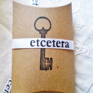 Peut inclure: Un sac cadeau en papier brun avec un ruban blanc et le mot "etceter" imprim&eacute; dessus. Le sac a un motif en forme de cl&eacute; imprim&eacute; sur le devant.