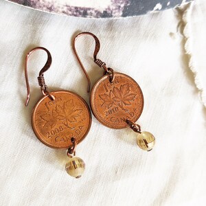 Peut inclure: Une paire de boucles d'oreilles de couleur cuivre avec des pi&egrave;ces de monnaie canadiennes de 1 cent de 2008 et 2010, chacune avec un motif de feuille d'&eacute;rable. Les pi&egrave;ces sont attach&eacute;es &agrave; des perles de verre transparentes.
